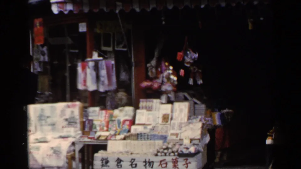 1950s Japan market stall display with stacked packages, hanging merchandise, Japanese signage, and a shadowed walkway beside it.