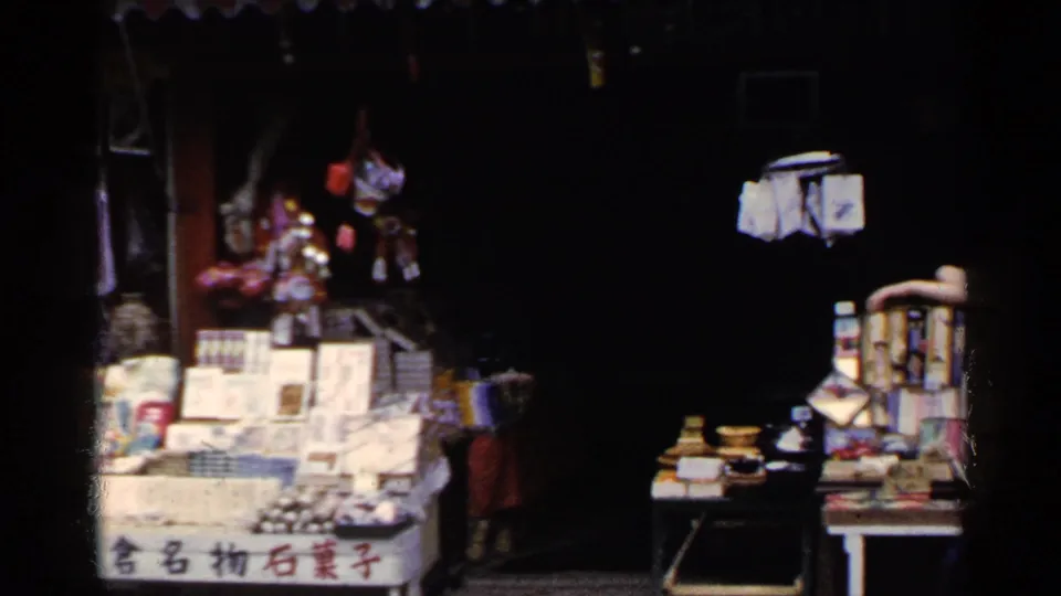 1950s Japan home-movie frame showing the market stall from a darker angle, with packaged goods, hanging items, and a small display table.