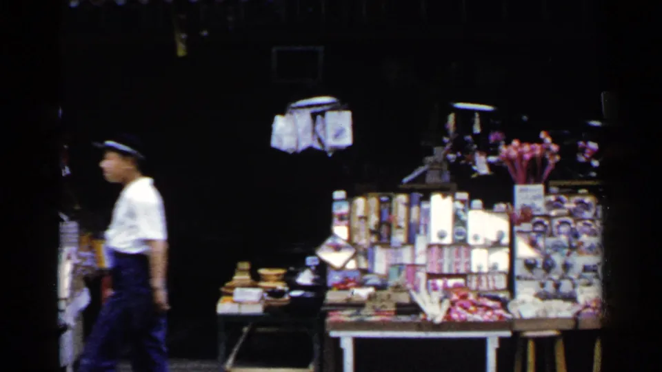 1950s Japan street-market frame with a white-shirted pedestrian passing display tables covered in small goods and packages.