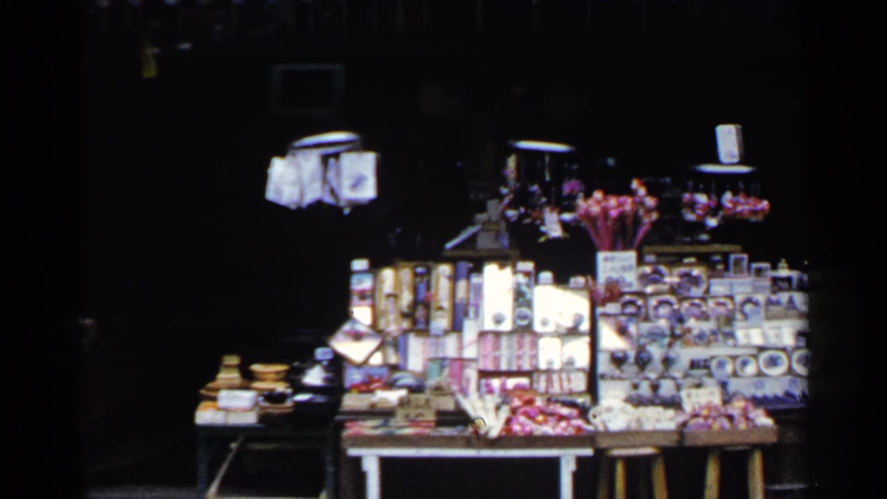 1953 Japan home-movie frame showing a souvenir stall with boxed goods, small figurines, and bright display items against a dark shopfront.