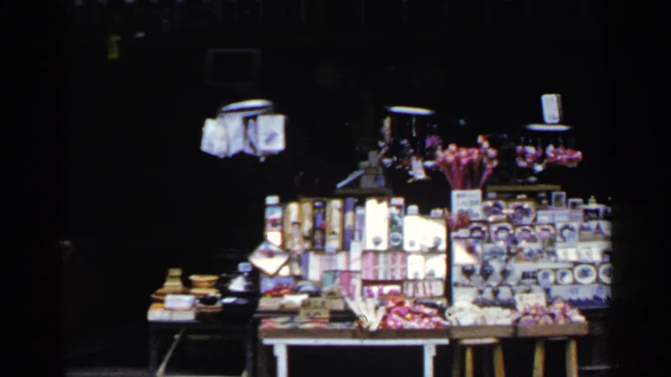 1953 Japan home-movie frame showing a souvenir stall with boxed goods, small figurines, and bright display items against a dark shopfront.