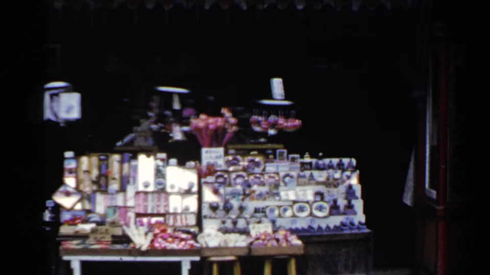The stall remains in view with rows of boxed goods, figurines, and small colorful souvenirs arranged on shelves and tables.