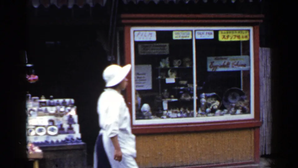 A woman in a white dress and wide-brimmed hat walks past a Japanese shop window with ceramics, glassware, signs, and a nearby souvenir display.