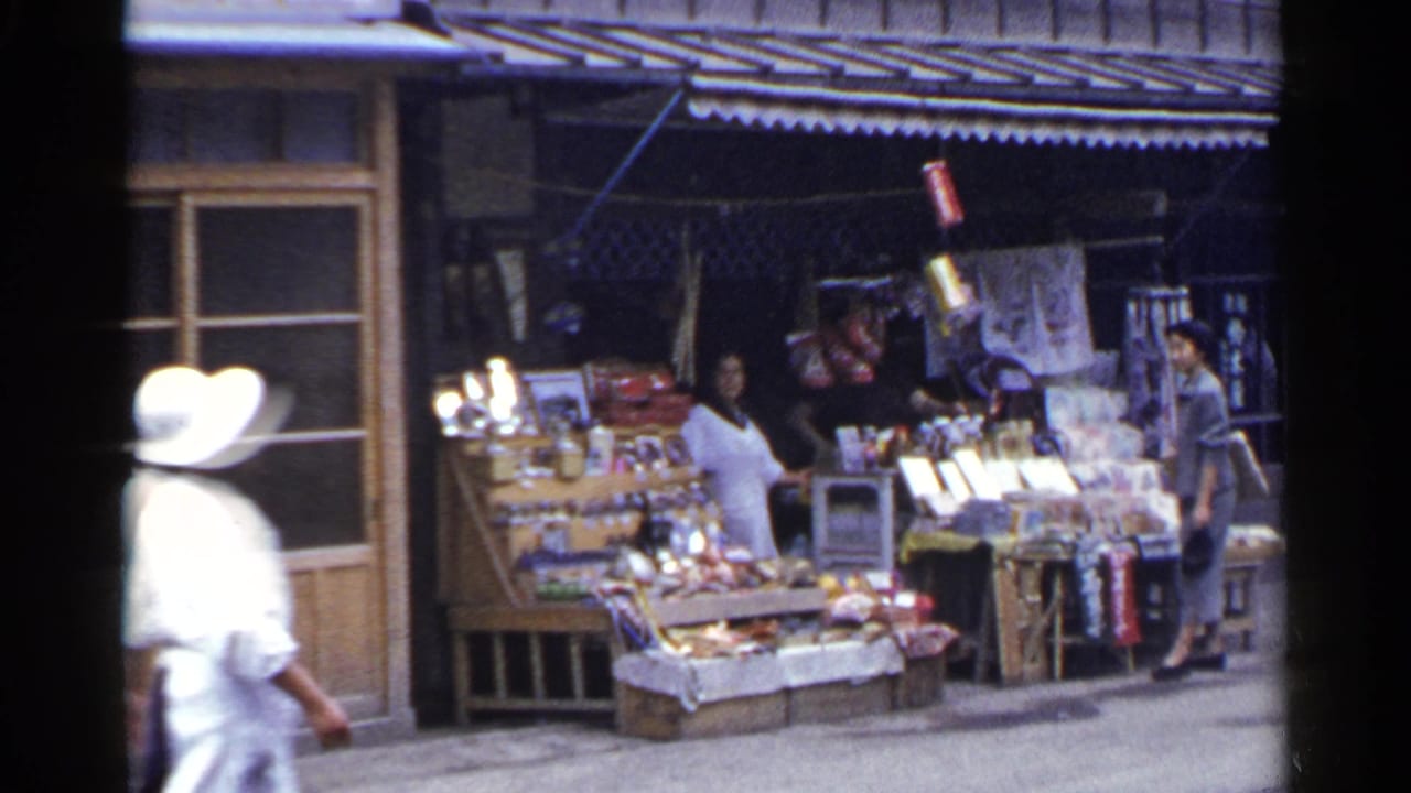 In 1953 Japan, a woman in a white hat walks past an open street stall with displayed goods and a wooden storefront.