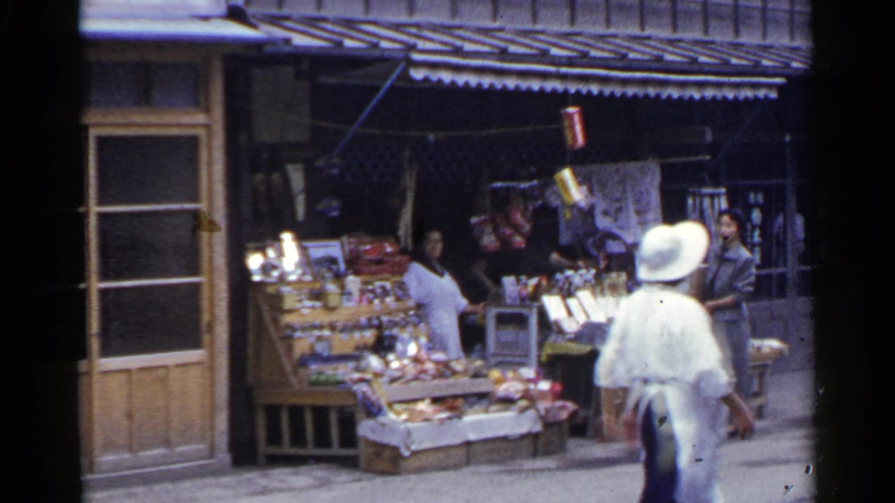 A woman in a light outfit and white sun hat passes a Japanese street vendor stall as another adult woman stands behind the display.