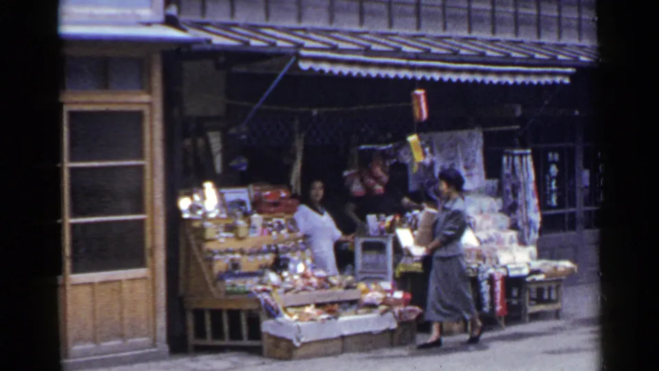 In the Japan street scene, a woman in a gray suit stands near a produce or goods display beneath a striped awning.
