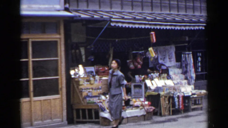 A woman in a gray suit walks past the street vendor stall in 1953 Japan, with open displays and hanging goods behind her.