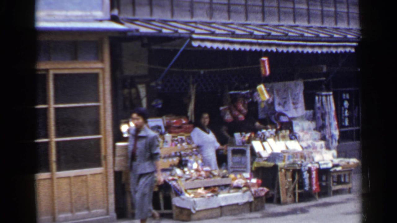 1953 Japan street scene with one woman at a produce stand and another woman passing beside crates under a storefront awning.