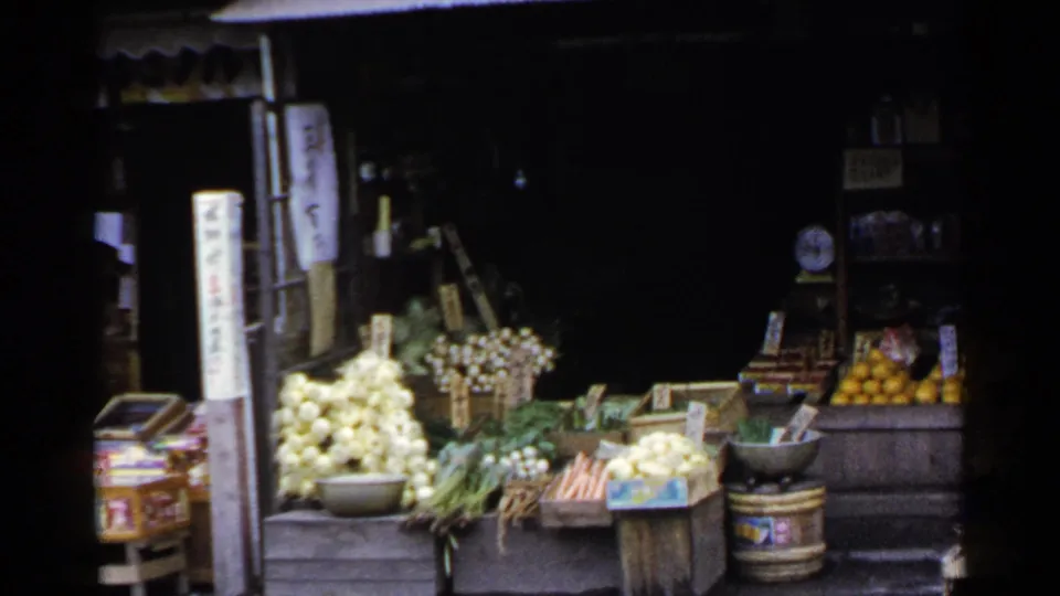 1953 Japan frame showing leafy vegetables, pale produce, fruit crates, and signs outside an open-front market shop.
