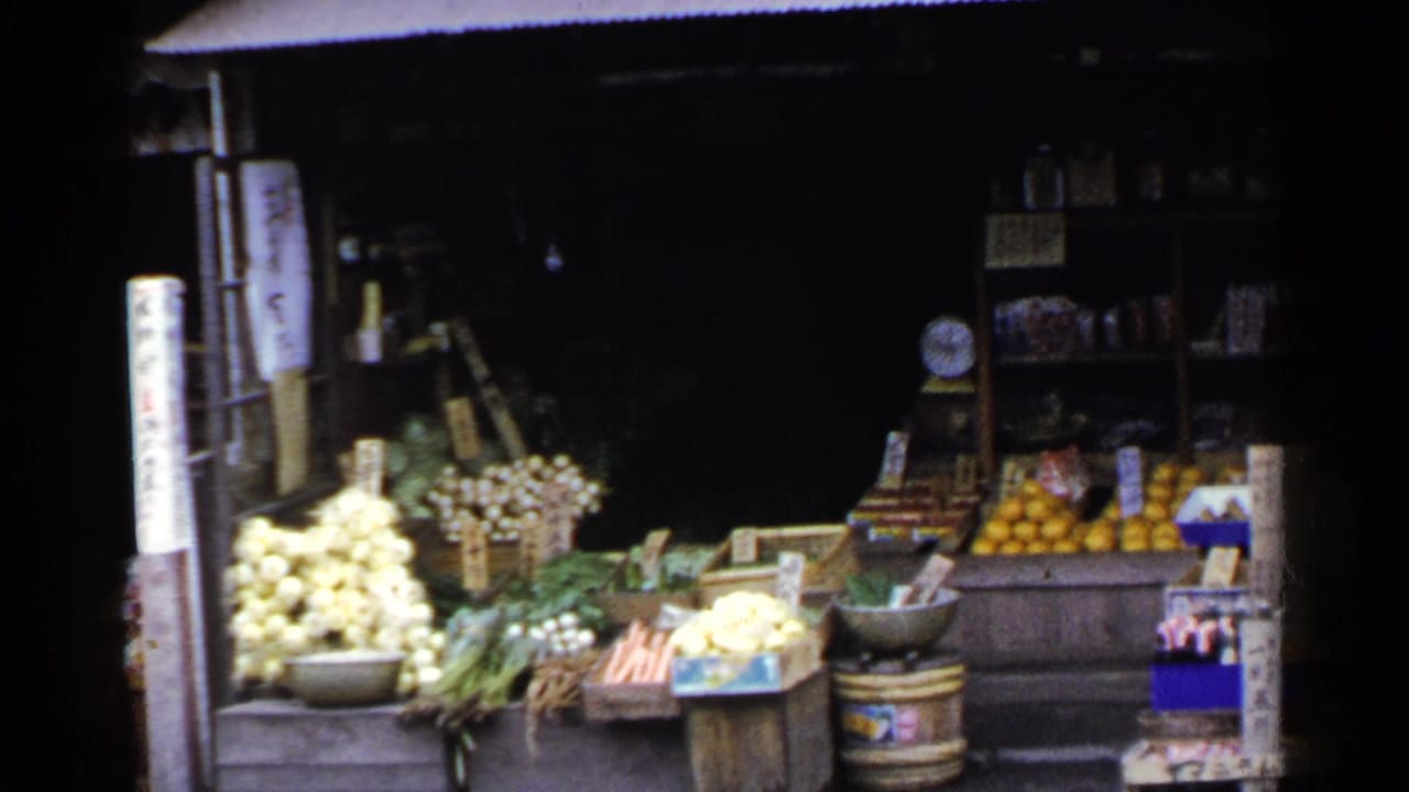 Produce display outside a dark storefront with onions, greens, carrots, oranges, bowls, and shelves.