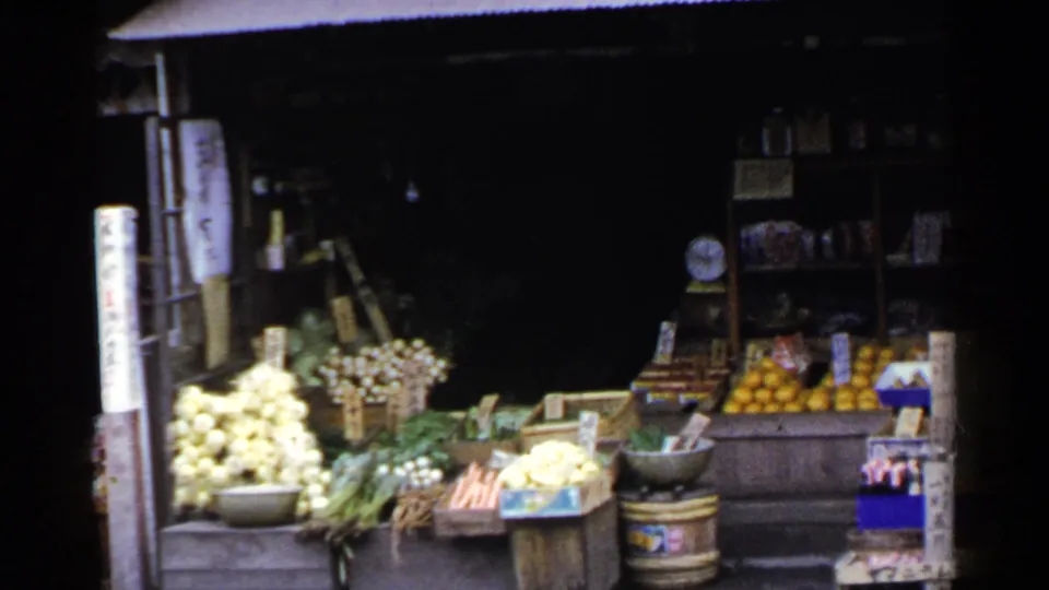 1953 Japan 8mm frame of a street produce display with piled onions, greens, carrots, oranges, display bowls, shelves, and Japanese signage.