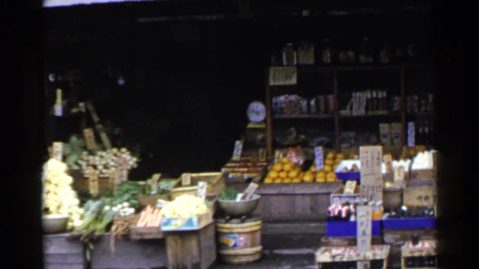 1953 Japan 8mm frame showing the produce stand from farther right, with oranges, white onions, vegetables, crates, boxed goods, and shelves.