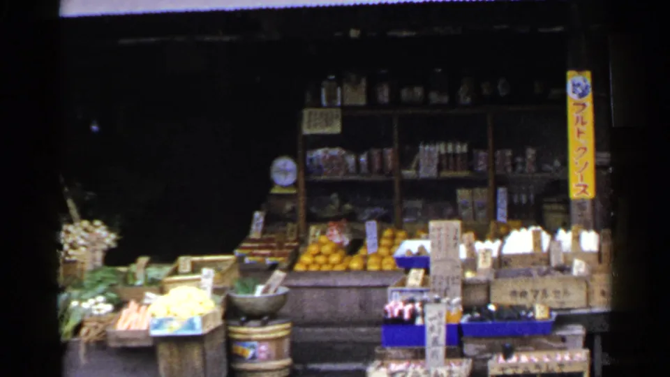 1953 Japan 8mm frame showing the right side of a produce storefront with oranges, onion piles, packaged goods, shelves, and a vertical yellow sign.