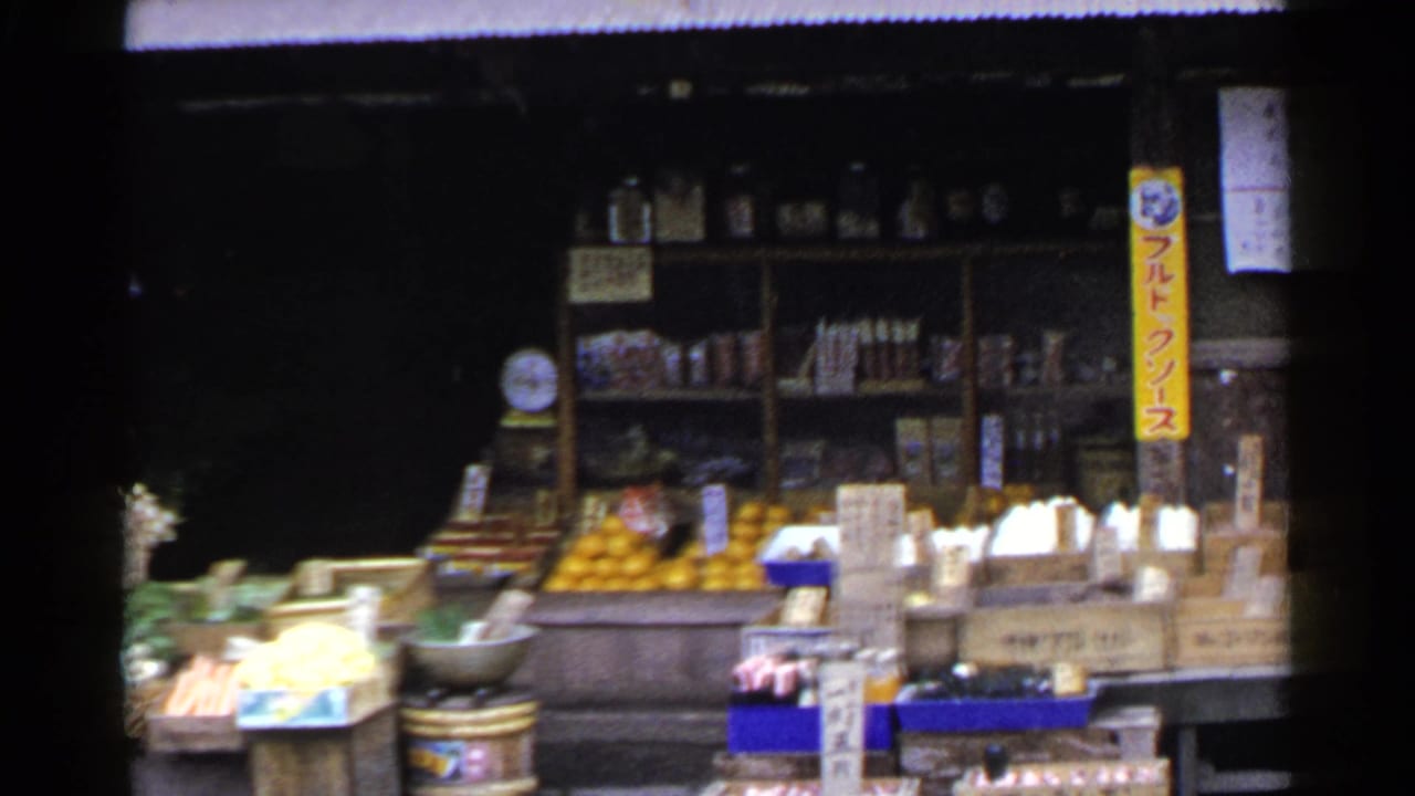 1953 Japan home-movie frame showing a wooden produce stand with oranges, boxed goods, Japanese signs, and bottles on rear shelves.