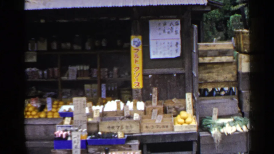 1953 Japan home-movie still showing crates of produce, oranges, leafy vegetables, Japanese price signs, shelves, and part of a wooden doorway.