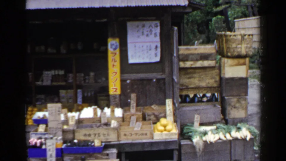 1953 Japan frame showing the produce stand from a slightly shifted angle, with oranges, greens, crates, signs, and greenery at the side.
