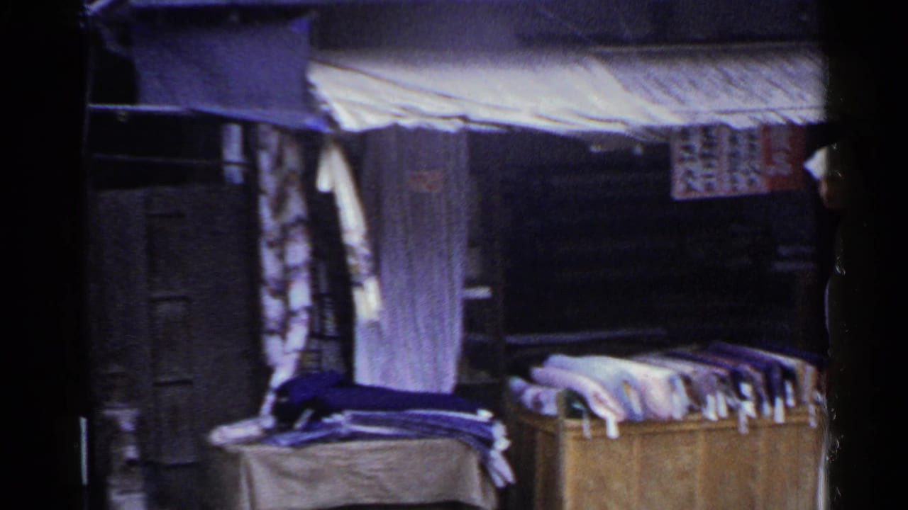 1953 Japan home-movie frame showing a market stall with folded fabric, hanging textiles, signage, and a shaded storefront.