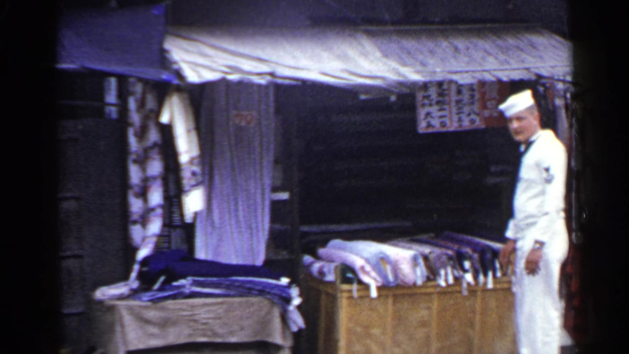 1953 Japan frame showing a U.S. Navy sailor smiling near folded textiles, hanging fabric, an awning, and visible Japanese signage.
