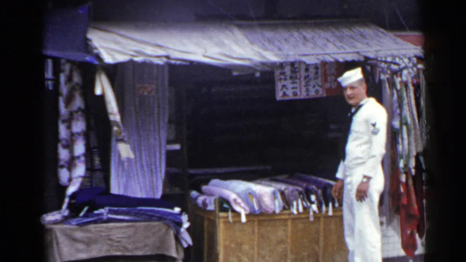 1953 Japan home-movie frame with a sailor in white uniform near a wooden counter covered with folded textiles and hanging cloth.