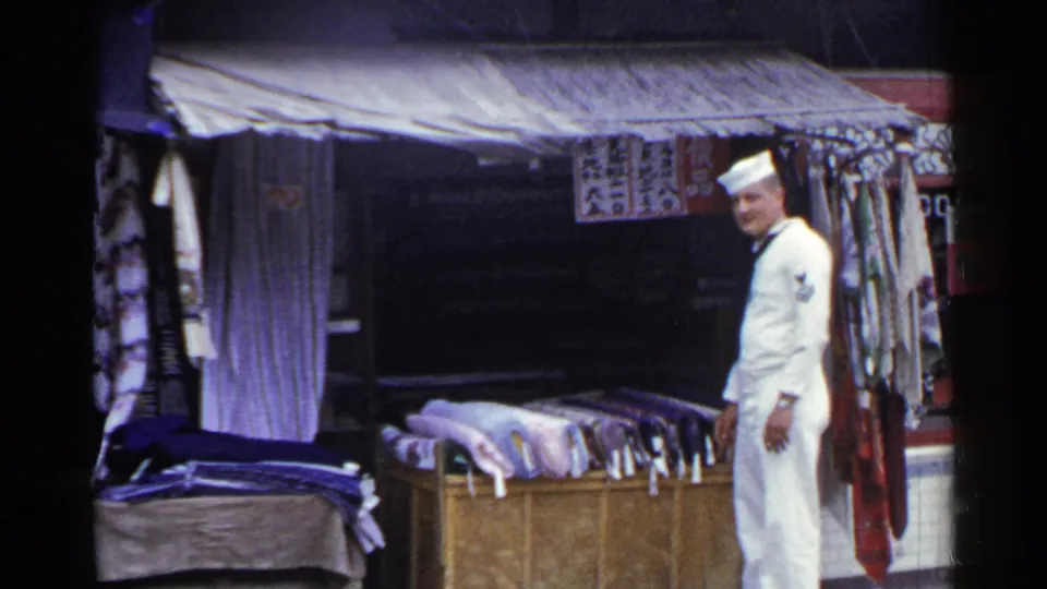 1953 Japan frame showing a smiling U.S. Navy sailor at a market stall with folded cloth, hanging fabric, awning, and signage.