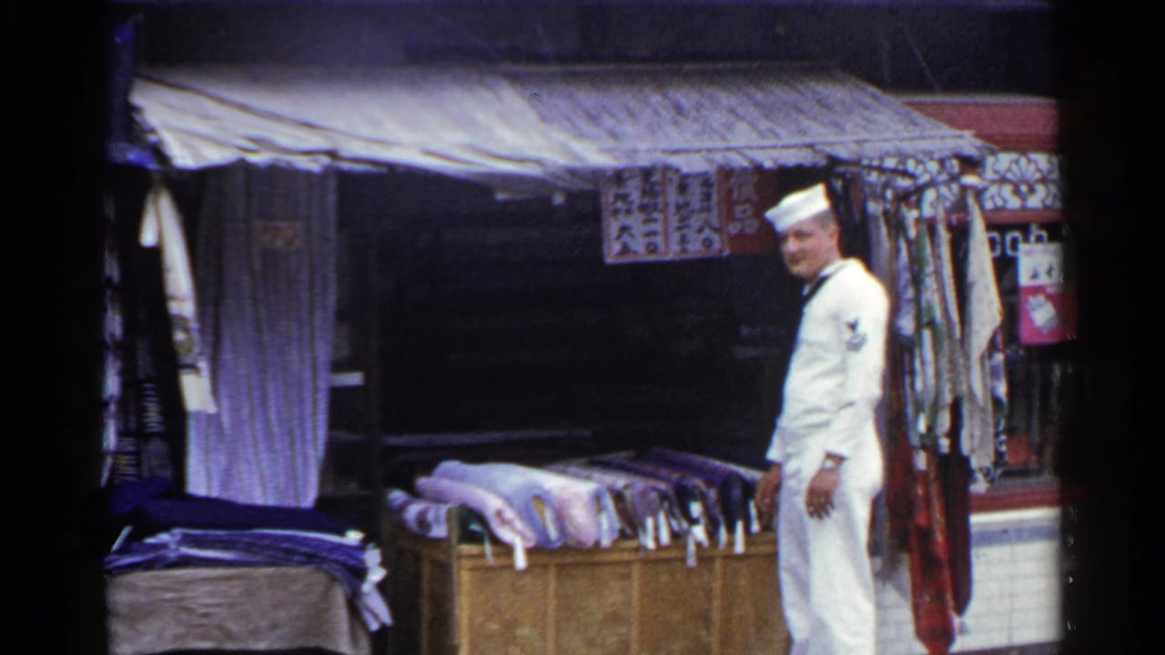 In 1953 Japan, a sailor in a white uniform stands near rolled rugs or textiles beneath a shop canopy with Japanese signage above.