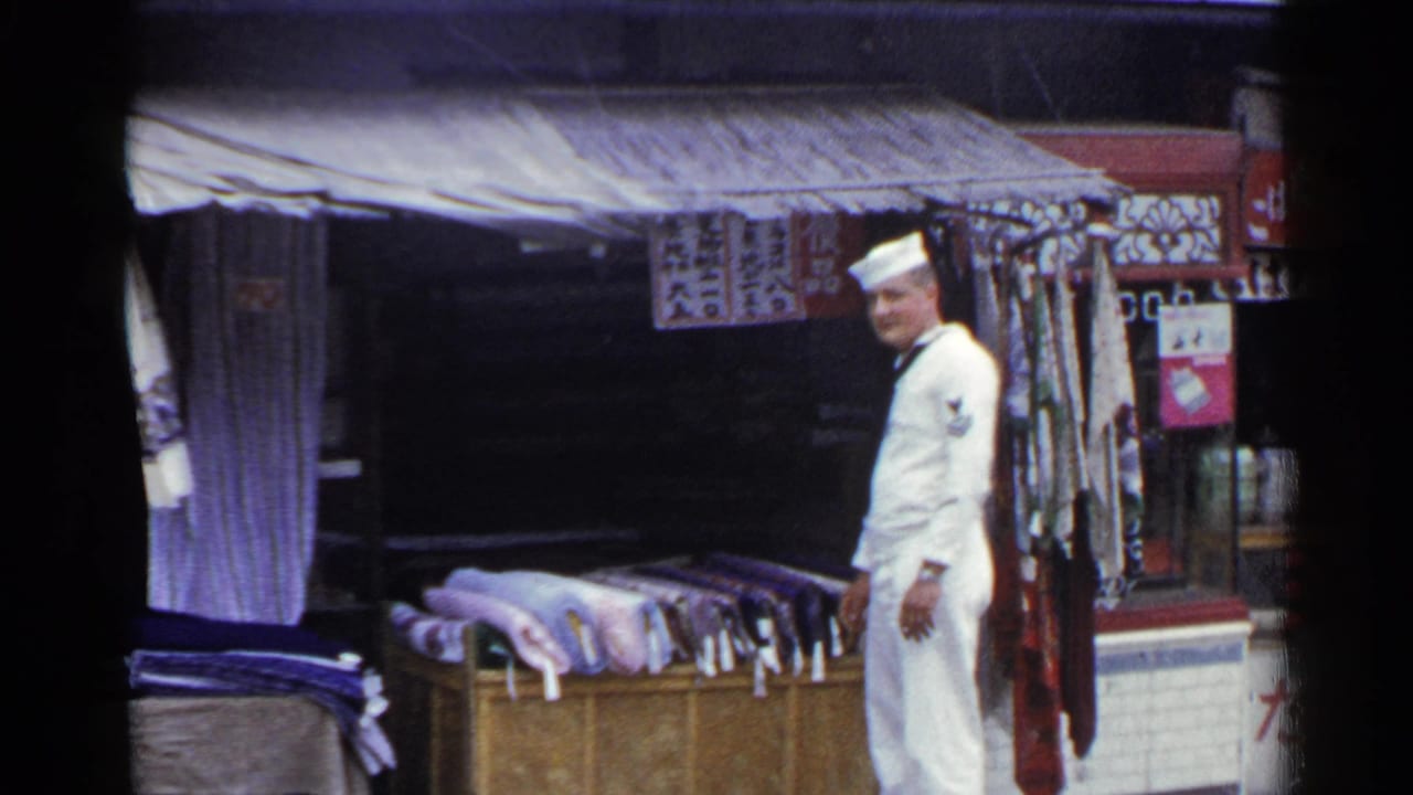 The sailor remains beside the outdoor textile display in Japan, with rugs, hanging fabric, and shop signs visible around the stall.