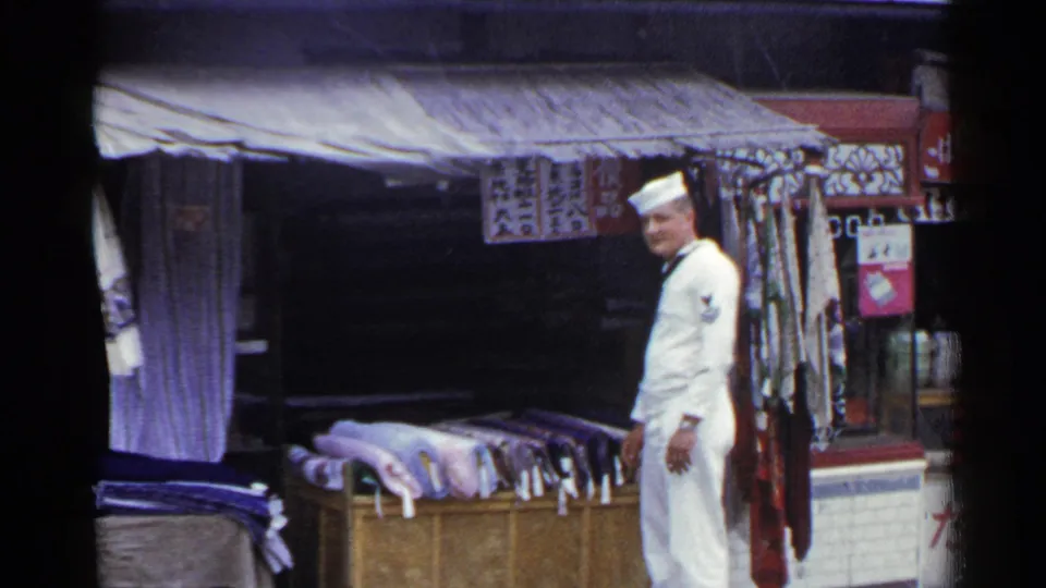 The sailor remains beside the outdoor textile display in Japan, with rugs, hanging fabric, and shop signs visible around the stall.