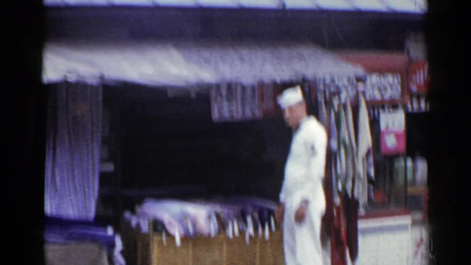 A soft, slightly blurred 8mm frame shows the sailor beside the textile stall, with the canopy, hanging fabric, and rolled goods still visible.