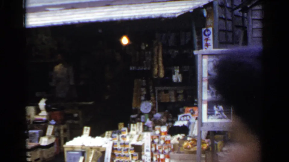 1953 Japan 8mm frame of a market stall with stacked goods and signage, briefly obscured by a person passing near the lens.