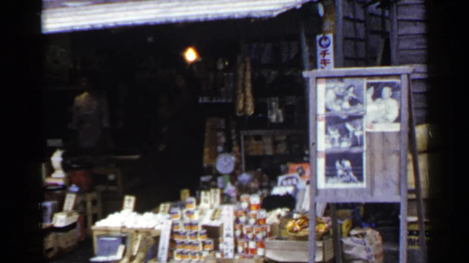 1953 Japan street-market frame showing a stocked food stall, vertical signage, photo display board, and warm light in the interior.