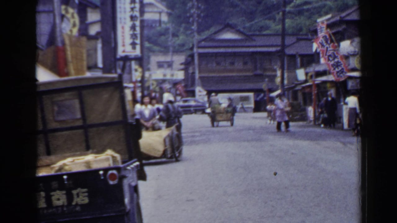 1953 Japan home-movie frame showing a town street with a handcart, pedestrians, Japanese signage, and wooden buildings.