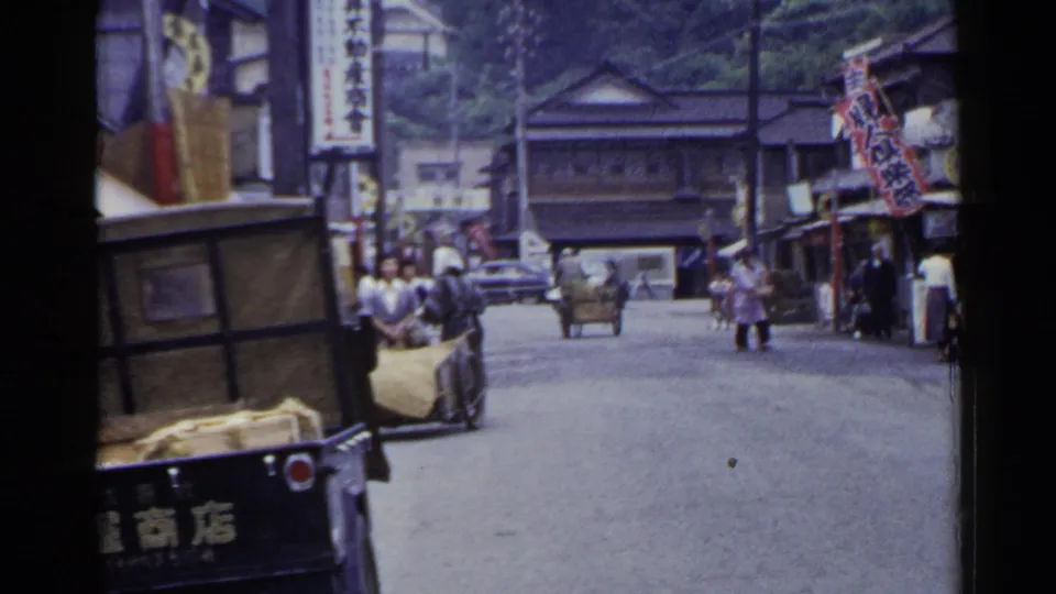 1953 Japan home-movie frame showing a town street with a handcart, pedestrians, Japanese signage, and wooden buildings.