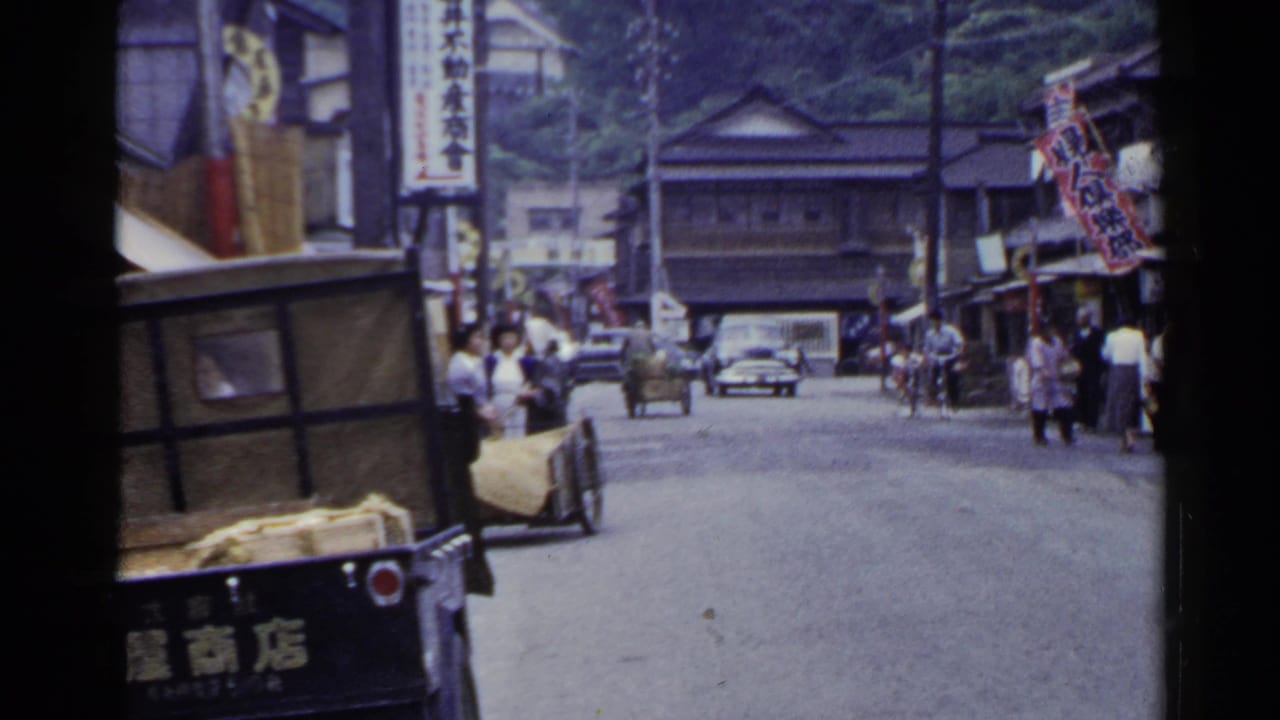1953 Japan street scene with pedestrians near storefronts as a cart and classic car appear along the road.
