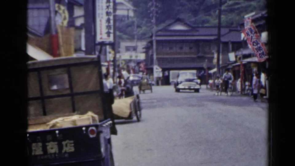 1953 Japan home-movie frame of a town road where a car approaches behind a cart, with pedestrians beside shopfronts.
