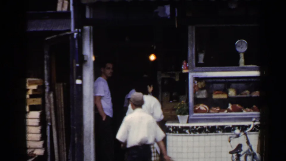 1953 Japan market entrance with customers near a tiled counter and display case, filmed from a handheld home-movie viewpoint.