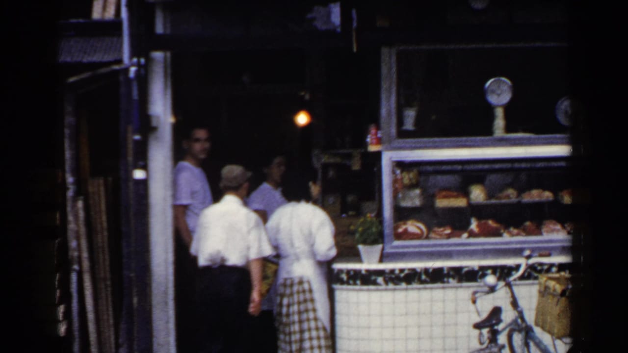 1953 Japan home-movie frame showing people gathered by a tiled deli counter, glass display case, and bicycle with basket.