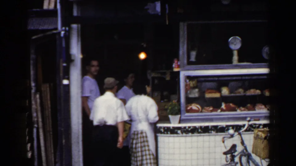 1953 Japan home-movie frame showing people gathered by a tiled deli counter, glass display case, and bicycle with basket.