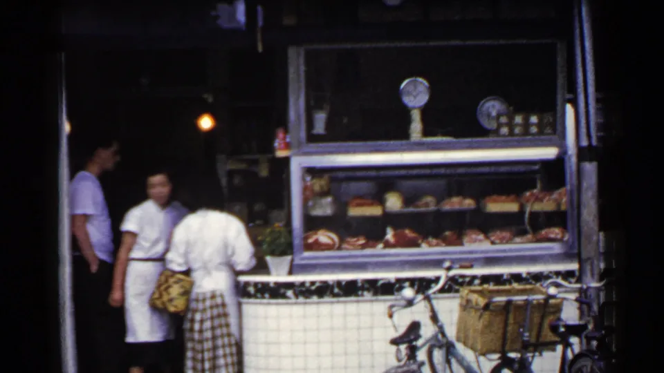 1953 Japan home-movie still with a worker in white, nearby customers, a glass deli case, tiled frontage, and parked bicycle.