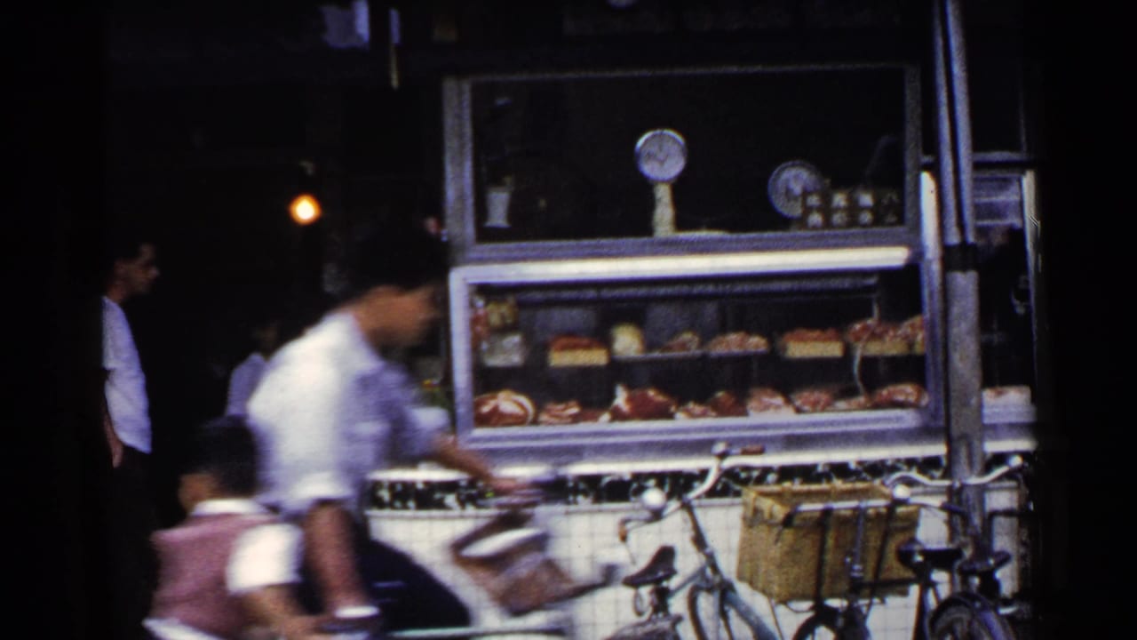 1953 Japan home-movie frame showing a tiled roadside food stall, displayed goods, bicycles, and a blurred passerby near the shopfront.