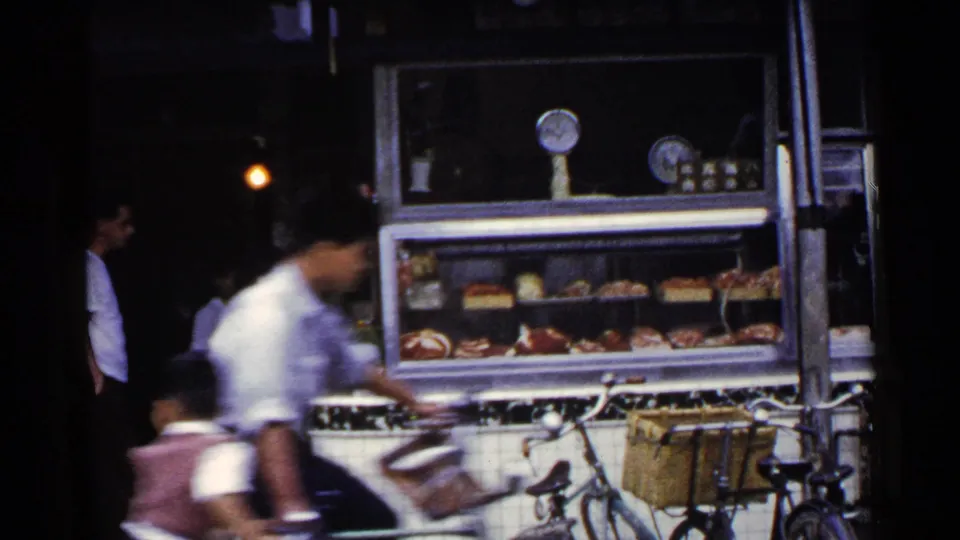 1953 Japan home-movie frame showing a tiled roadside food stall, displayed goods, bicycles, and a blurred passerby near the shopfront.