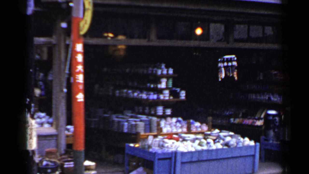 1953 Japan market view with stocked shelves, a blue display bin of goods, dim shop lighting, and a red pole with Japanese characters.
