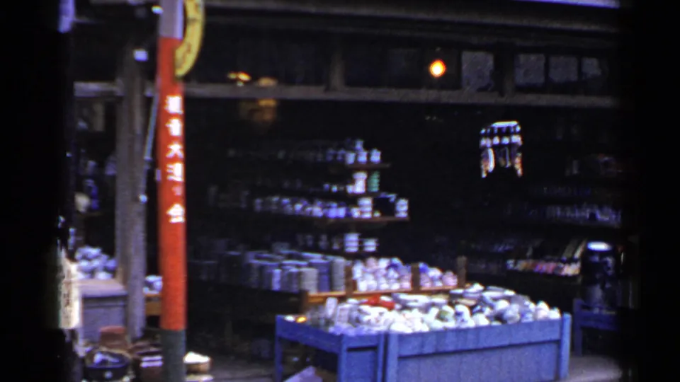 1953 Japan market view with stocked shelves, a blue display bin of goods, dim shop lighting, and a red pole with Japanese characters.