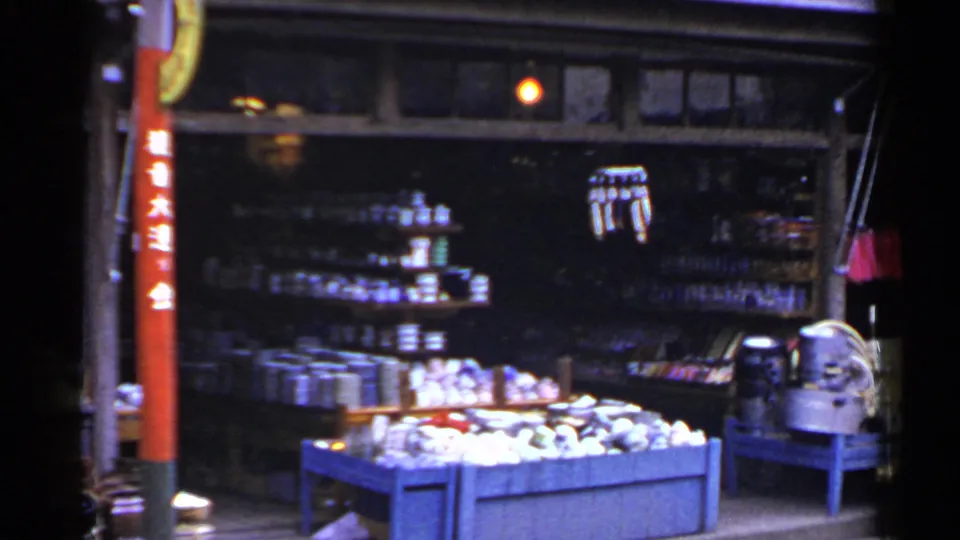 1953 Japan home-movie frame of a roadside market display with stacked goods, blue produce bin, red sign pole, and dark storefront shelves.