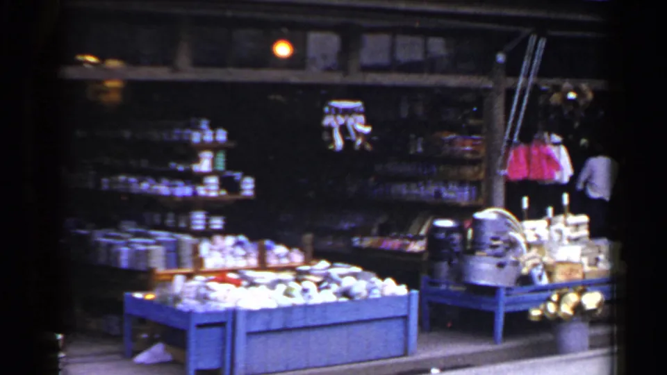 1953 Japan street-market frame showing stocked shelves, a blue bin of goods, hanging items, and a person near the shop edge.