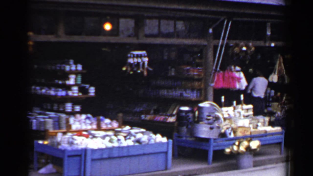 1953 Japan home-movie frame showing a market stall with blue display tables, goods for sale, hanging pink and white items, and a man inside.