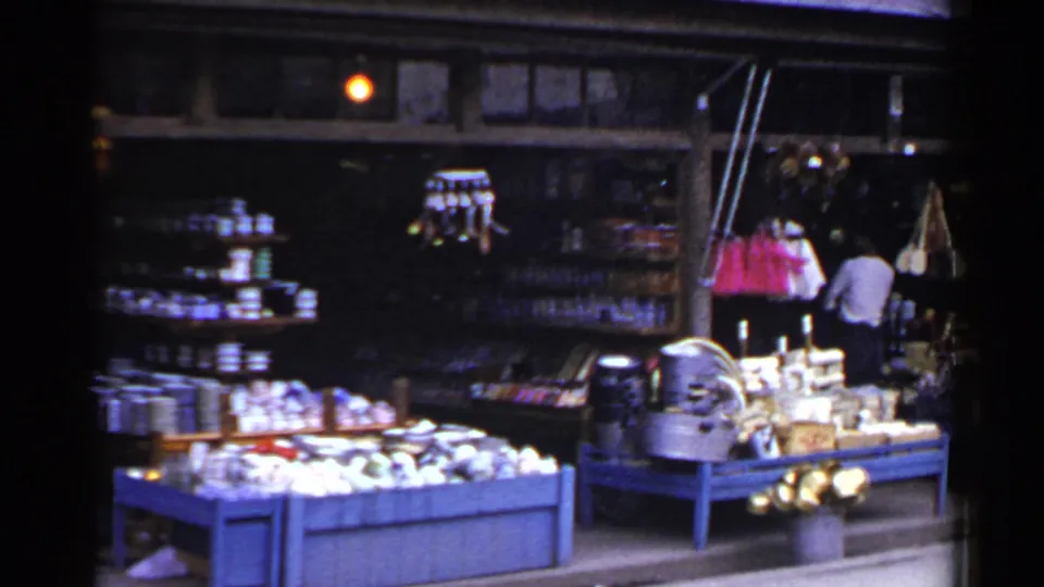 1953 Japan home-movie frame showing a market stall with blue display tables, goods for sale, hanging pink and white items, and a man inside.