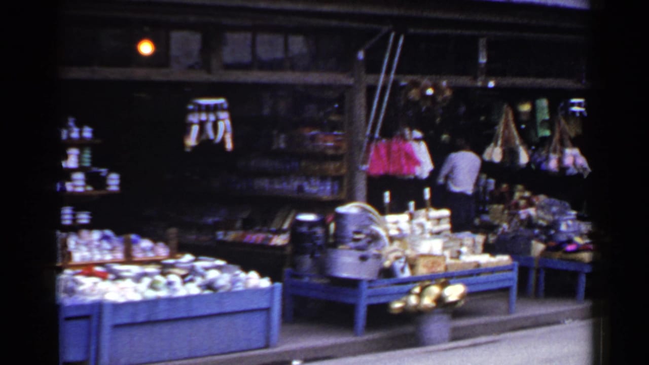 Market display with blue tables, baskets or bags, hanging goods, and a man standing at the back.