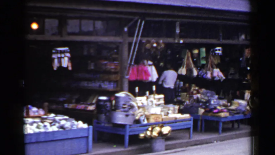 1953 Japan home-movie still with a blue-painted market display, small goods, hanging items, yellow flowers, and a man inside the stall.