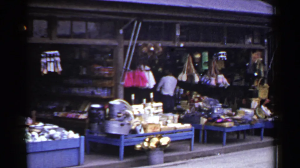 1953 Japan market frame showing clustered goods on blue tables, hanging bags or garments, yellow flowers near the entrance, and a man inside.