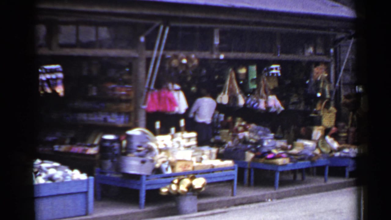 1953 Japan 8mm frame showing a market storefront with blue tables, displayed goods, baskets, and people browsing near the entrance.